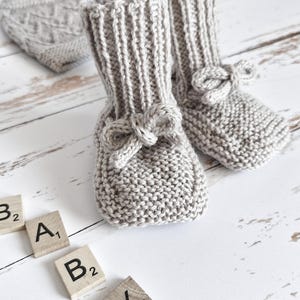 Peut inclure: Une paire de chaussons en tricot pour bébé avec un détail en nœud de couleur beige clair. Les chaussons sont sur une surface en bois blanc avec les lettres "BABY" écrites en carreaux de bois.