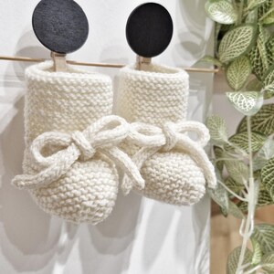 Peut inclure: Une paire de chaussons de bébé en tricot ivoire, avec des détails de nœuds. Les chaussons sont suspendus à une tige en bois par des pinces à linge. Le fond est un mur blanc avec une plante décorative.