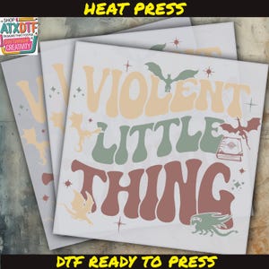 May include: A white heat transfer design with the text "VIOLENT LITTLE THING" in a retro font. The design features several stylized dragons and stars. The text "DTF READY TO PRESS" is at the bottom of the design.