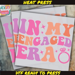 May include: A pink, orange, and purple iron-on design that reads "In My Engaged Era" with a diamond ring graphic. The design is ready to be pressed onto a t-shirt or other fabric.