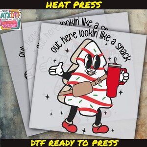 May include: A white and red candy cane Christmas tree character with googly eyes and a red thermos. The character is wearing a brown belt and has a white background with stars. The text "out here lookin' like a snack" is written in black above the character. The text "DTF READY TO PRESS" is written in black at the bottom of the image.