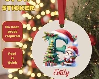 May include: A round Christmas ornament with a red ribbon. The ornament features a green letter "E" decorated with a Christmas tree and Santa Claus. The name "Emily" is written in red script. The text "UV DTF STICKER" and "Peel & Stick" are also visible.