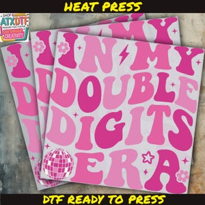 May include: Pink and white DTF (Direct to Film) heat transfer sheets with the text "IN MY DOUBLE DIGITS ERA" in a groovy font. The design includes a disco ball, flowers, and stars. The words "HEAT PRESS" and "DTF READY TO PRESS" are also visible.