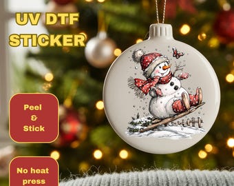 May include: A round, off-white Christmas ornament featuring a snowman wearing a red and white hat, scarf, and boots, skiing down a snowy hill. The ornament has the text "UV DTF STICKER" and "Peel & Stick" on the left side.