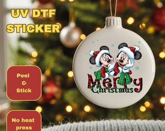 May include: A white Christmas ornament featuring Mickey and Minnie Mouse in Santa hats and scarves, with the words "Merry Christmas" in a plaid design. The image also includes text that says "UV DTF STICKER", "Peel & Stick", and "No heat press required."