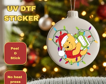 May include: A round, off-white Christmas ornament featuring Winnie the Pooh in a Santa hat, holding a gift. The ornament is decorated with colorful Christmas lights. Text on the image reads "UV DTF STICKER", "Peel & Stick", and "No heat press required."