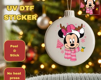 May include: A white Christmas ornament featuring Minnie Mouse in a pink hat, scarf, and antlers. The ornament has a pink Christmas tree and snowflakes. The image also includes text that says "UV DTF STICKER", "Peel & Stick", and "No heat press required."