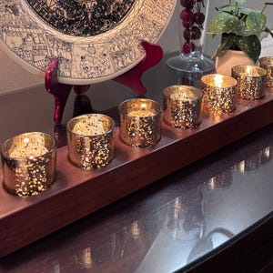 May include: A wooden candle holder with seven gold speckled votive candle holders, each containing a lit candle. The holder sits on a dark surface, with a decorative plate and other items in the background. The candles provide a warm, inviting glow.