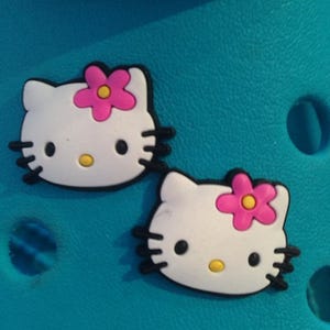 May include: Two white, cartoon cat-shaped shoe charms with black outlines, whiskers, and eyes, and a pink flower on their heads. The charms have yellow noses and are set against a turquoise background.