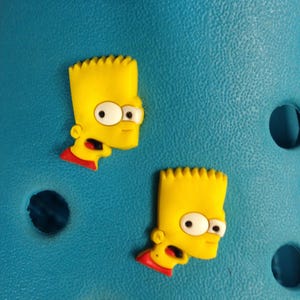 May include: Two yellow Bart Simpson shoe charms with black and white eyes and red accents. The charms are on a textured teal surface with dark blue circular holes.