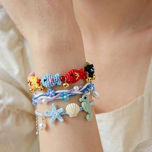 Op de afbeelding: Een set van drie gelaagde armbanden met kleurrijke gehaakte vissen, kleine gouden belletjes, witte parels, blauw gevlochten koord en lichtblauwe gehaakte bedels in de vorm van een zeester, een schelp en een zeepaardje met kleine blauwe bloemen.