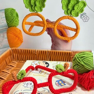 May include: Two pairs of novelty glasses made from crocheted yarn. One pair is orange with green leafy accents resembling carrots, and the other pair is red with small green and brown stem details resembling apples, both featuring round frames.