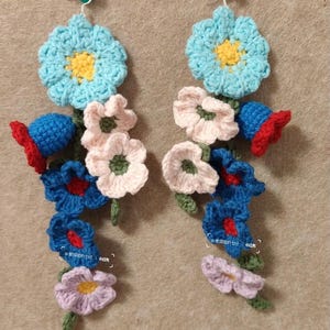 Pode incluir: Um par de brincos de crochê feitos à mão com um design floral. Cada brinco tem uma grande flor azul clara com um centro amarelo e uma cascata de flores menores em rosa, azul e roxo. Os brincos são presos a um alfinete verde.