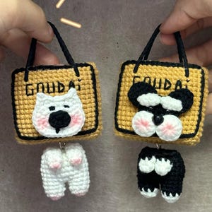 Puede incluir: Dos colgantes de bolso de perro hechos a mano con ganchillo. Cada colgante presenta un cuadrado amarillo con la palabra "GOUDA" y una cara de perro. Un perro es blanco con detalles en rosa, el otro es blanco y negro. Ambos tienen asas negras y traseros de perro colgantes.