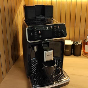 Peut inclure: Une machine à expresso Philips noire avec une garniture argentée et une tasse noire. La machine dispose d'un écran numérique et d'un réservoir d'eau sur le dessus. La machine est sur une surface en bois.