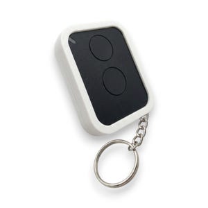 Peut inclure: Une télécommande rectangulaire avec un cadre extérieur blanc et un panneau central noir comportant deux boutons circulaires. Un petit anneau porte-clés en métal argenté est fixé au bas de l'appareil par une courte chaîne, créant un design pratique.