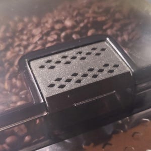 May include: Close-up of a black plastic coffee bean container lid with a rectangular, perforated vent. The lid is set against a backdrop of brown coffee beans, suggesting a storage container for coffee beans.