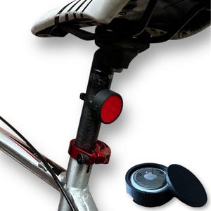 以下が含まれることがあります： 黒と赤の自転車用リフレクターと黒のAirTagホルダーが表示されています。リフレクターは黒いシートポストに取り付けられています。AirTagホルダーは黒色で、取り外し可能な蓋が付いています。自転車のサドルは白と黒です。
