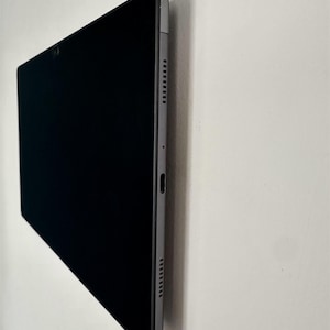 May include: A black tablet with a gray frame is mounted on a white wall. The tablet's screen is dark, and the side profile shows speaker holes and a charging port. The text "Support invisible" is at the bottom.