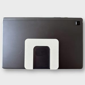May include: A dark gray tablet with a white, U-shaped stand attached to the back. The stand is made of a textured material. The tablet has a camera on the top right corner.