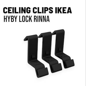 Op de afbeelding: Drie zwarte plafondclips met de tekst "CEILING CLIPS IKEA HYBY LOCK RINNA" op een witte achtergrond. De clips zijn gemaakt van mat materiaal en hebben een haakachtig ontwerp.