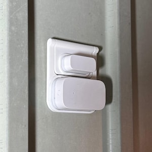 Puede incluir: Un dispositivo domótico blanco, de forma rectangular, montado en una pared. El dispositivo tiene dos componentes rectangulares, uno encima del otro. La pared es de color gris claro.