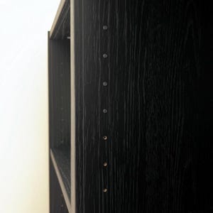 K&ouml;nnte beinhalten: Nahaufnahme eines schwarzen B&uuml;cherregals aus Holz mit sichtbarer Holzmaserung. Das B&uuml;cherregal hat mehrere Regale und ist aus dunklem Holz gefertigt. Kleine Metallbefestigungen sind an der Seite des B&uuml;cherregals sichtbar.