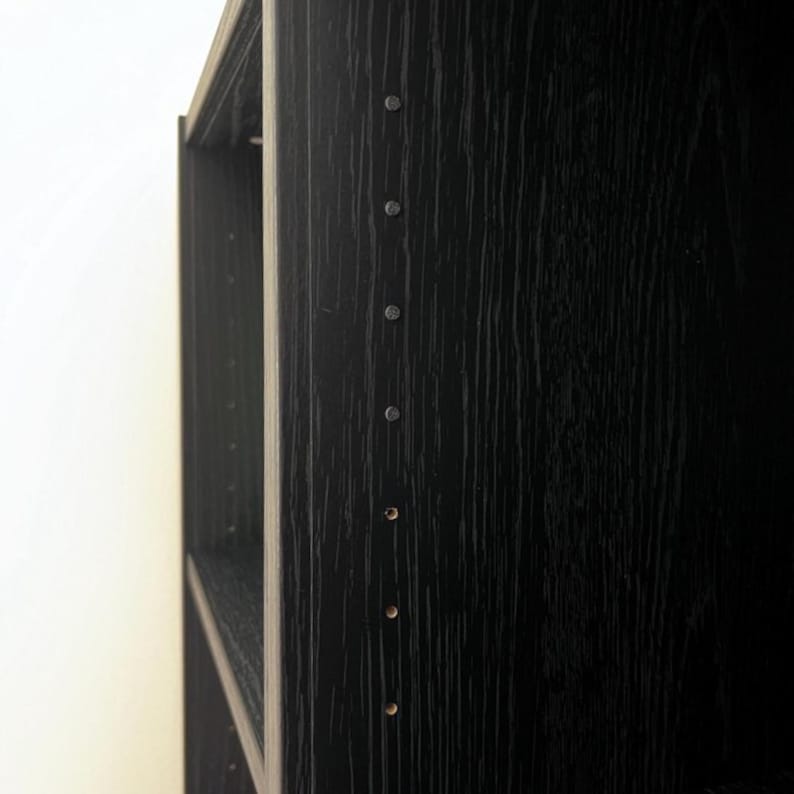 K&ouml;nnte beinhalten: Nahaufnahme eines schwarzen B&uuml;cherregals aus Holz mit sichtbarer Holzmaserung. Das B&uuml;cherregal hat mehrere Regale und ist aus dunklem Holz gefertigt. Kleine Metallbefestigungen sind an der Seite des B&uuml;cherregals sichtbar.