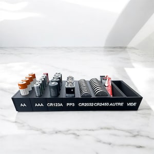 Puede incluir: Un organizador de baterías negro con compartimentos etiquetados para varios tamaños de batería, incluyendo AA, AAA, CR123A y PP3. El organizador contiene diferentes tipos de baterías, algunas visibles en naranja, plata y negro.