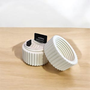 May include: A white, fluted, ceramic container with a removable lid, next to a matching lid. A black MicroSD adapter is inserted into the container. The container sits on a light wood surface.