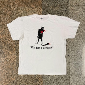 Könnte beinhalten: Weißes T-Shirt mit einer Grafik eines schwarzen Ritters mit Schwert, rotem Blutspritzer und dem Text "'Tis but a scratch!". Dem Ritter fehlt ein Arm. Das Shirt hat einen Rundhalsausschnitt und kurze Ärmel.