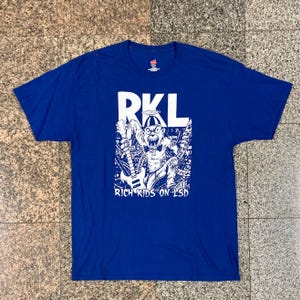 May include: Royal blue t-shirt with a white graphic. The graphic features the letters "RKL" above an illustration of a cartoon character playing a guitar, with the text "RICH KIDS ON LSD" below. The shirt is made of a soft material.