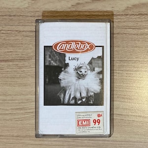 以下が含まれることがあります： Candleboxのアルバム「Lucy」のヴィンテージカセットテープ。カバーには、マスクとフリルカラーを身に着けた人物の白黒写真が写っています。テープは透明なプラスチックケースに入っています。