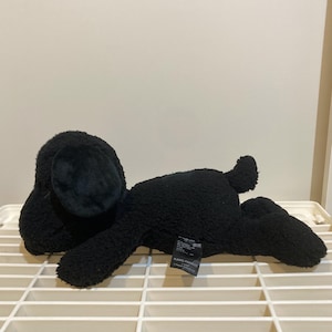 May include: A black plush dog toy lying down. The toy has large, floppy ears and a small tail. The plush is made of a soft, textured material. A tag is attached to the toy.