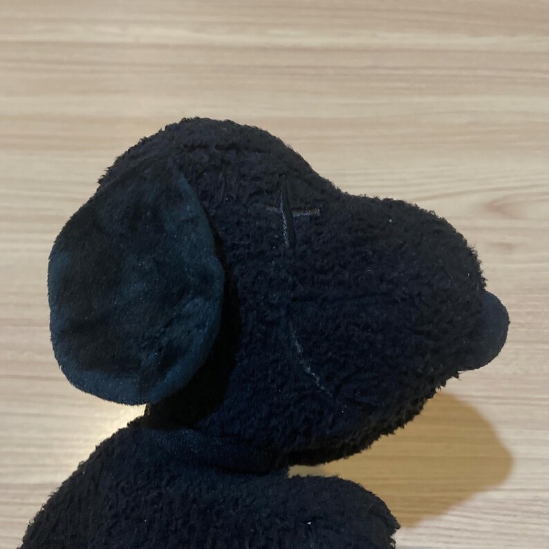 May include: A black plush dog toy, featuring a soft, textured exterior. The toy has large, floppy ears and a small, rounded nose. The dog's head is turned to the side, showcasing its profile. The toy is designed to resemble a cartoon dog.