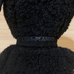 May include: Close-up of a black plush toy with the text "KAWS PEANUTS" embroidered on the neck. The toy has a textured, fuzzy appearance. The image focuses on the upper body of the toy.