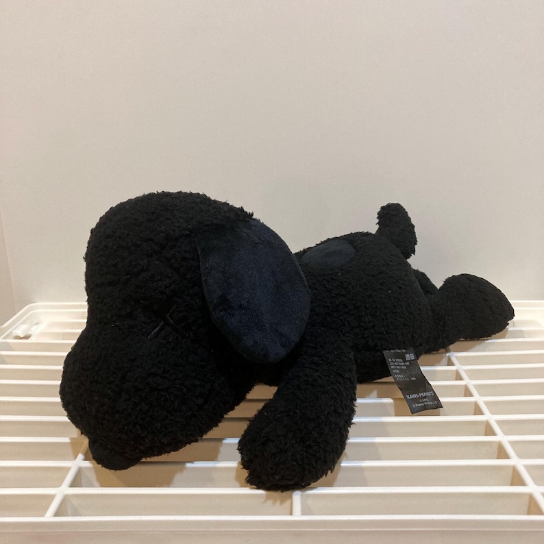 May include: A black plush dog toy with floppy ears and a stitched mouth. The dog is lying down on a white grid surface. The toy has a soft, textured appearance and a small tag attached to its side.