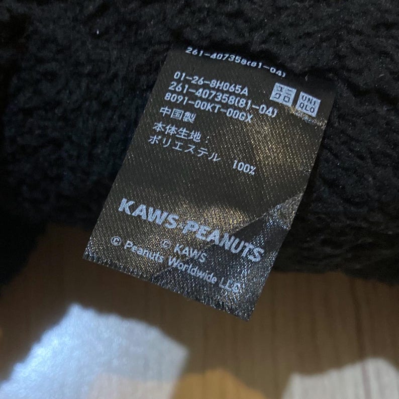 May include: A black tag with white text, featuring the KAWS PEANUTS logo and product information. The tag includes the text "Polyester 100%" and the UNIQLO logo.