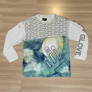 May include: White long-sleeve shirt with a graphic design featuring a hand outline over a yellow and blue background. The sleeves have the word "GLOVE" printed vertically. The shirt has the words "GLOVE BODY" printed repeatedly.
