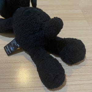 May include: A black plush toy of a cartoon character, lying on its stomach. The toy has a textured, fuzzy appearance. A tag with Japanese text is attached to the toy. The toy is on a light brown surface.