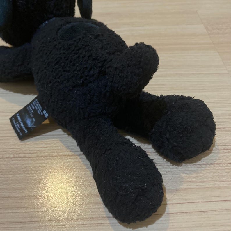 May include: A black plush toy of a cartoon character, lying on its stomach. The toy has a textured, fuzzy appearance. A tag with Japanese text is attached to the toy. The toy is on a light brown surface.