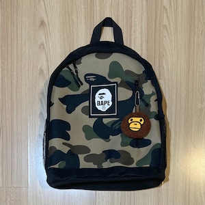May include: A camouflage-patterned backpack with black accents. The backpack features a black top handle, a front zipper pocket, and a small, round, brown keychain. The BAPE logo is displayed on a white square patch.