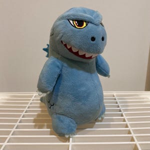 May include: A light blue plush Godzilla toy with a cartoonish design. The toy has a wide grin with white teeth, yellow and red eyes, and small dorsal fins. It is standing on a white wire shelf.