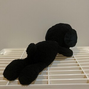 May include: A black plush toy dog lying on a white slatted surface. The dog has floppy ears and a small tail. The toy is made of a soft, textured material.