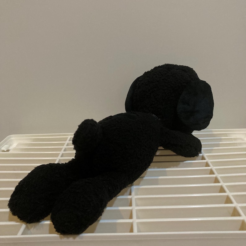 May include: A black plush toy dog lying on a white slatted surface. The dog has floppy ears and a small tail. The toy is made of a soft, textured material.