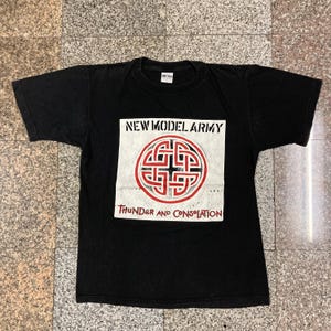 Pode incluir: Camiseta preta com um gráfico quadrado branco que apresenta a banda "New Model Army" e o título do álbum "Thunder and Consolation". O gráfico também inclui um design de nó celta vermelho.