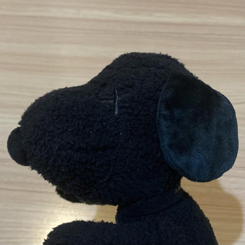 May include: A black plush dog toy, viewed in profile, with a prominent ear and a small, stitched detail above the eye. The toy has a soft, textured appearance and is set against a light-colored wooden surface.