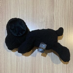 May include: A black plush dog toy with a textured, fuzzy exterior. The dog has large, floppy ears with a velvety interior. A small tag is attached to the dog's body. The toy is lying on a light-colored wooden surface.