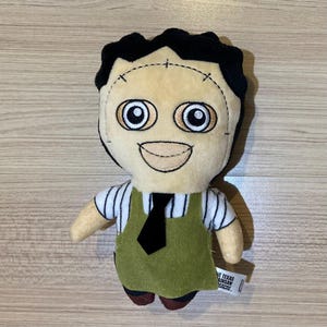 May include: Plush doll of a character with a stitched face, black hair, and large eyes. The doll wears a white striped shirt, green apron, and black tie. The doll is a character from The Texas Chainsaw Massacre.