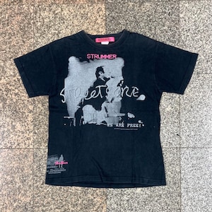 Może przedstawiać: Czarny t-shirt z szarą grafiką muzyka grającego na gitarze, z napisem "STRUMMER" powyżej i "Street One" poniżej. Koszulka ma również napis "WE ARE FREE!" i różową metkę z napisem "RALEIGH".